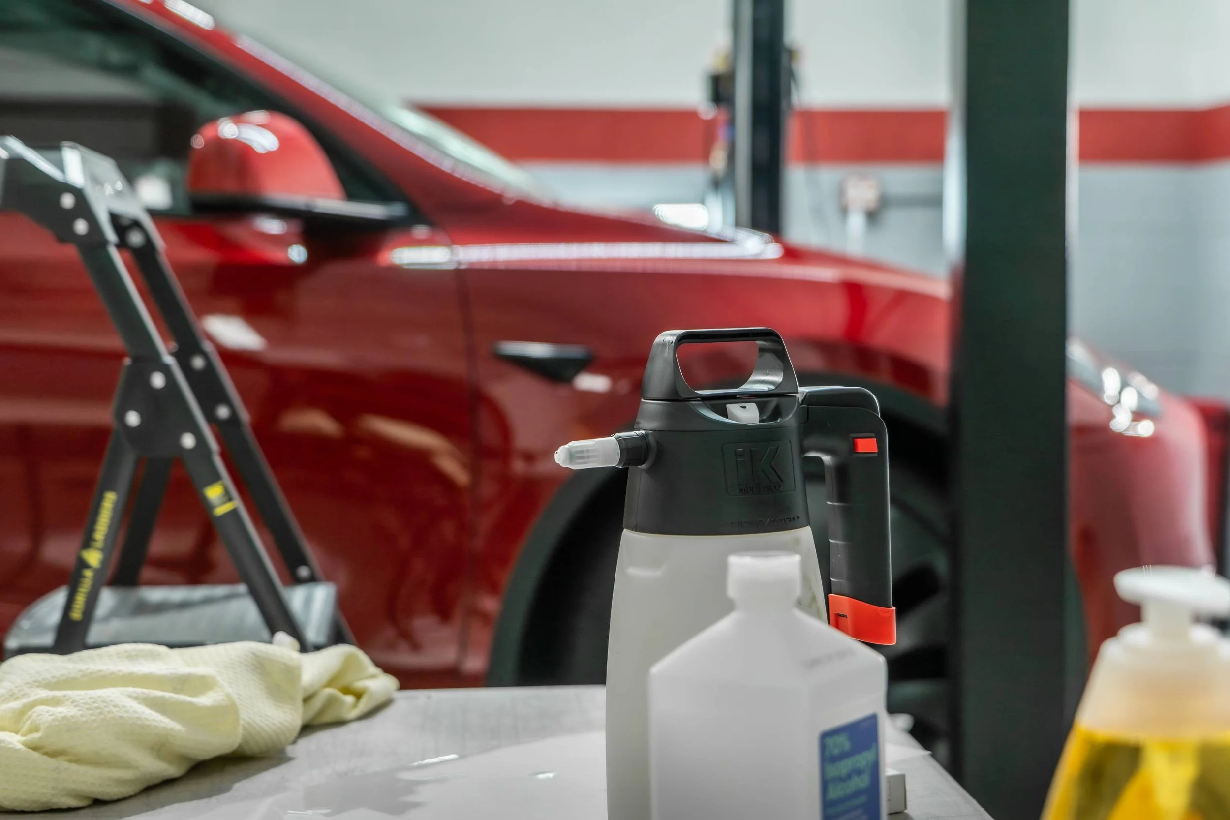 Cleaning and detailing supplies on a table in front of a red vehicle, including a spray bottle, a cleaning solution container, a microfiber cloth, and a steam cleaner in a garage.