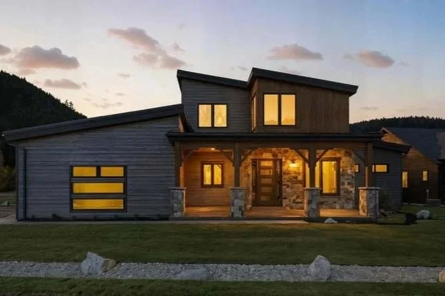 Modern two-story house with large windows and a front porch, illuminated at dusk with a mountain backdrop.