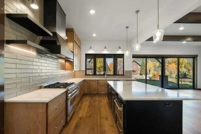 Modern kitchen with wooden cabinets, white countertops, black appliances, brick backsplash, large windows showing trees outside, and pendant lighting.