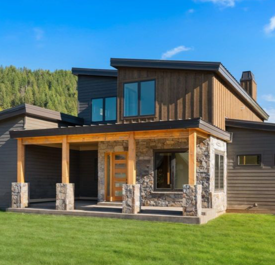 Modern two-story house with a mix of wooden and stone exterior, large windows, and a covered porch with wooden support beams, situated on a grassy lawn with a background of trees and a clear blue sky.
