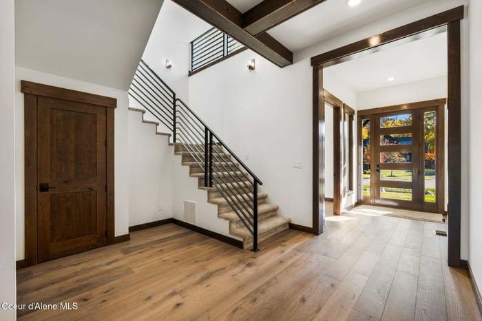 Interior view of a modern home entryway with wood flooring, a staircase with black metal railings, a wooden door, and large windows with sunlight coming through.