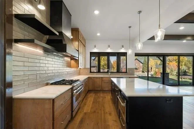 Modern kitchen with wooden cabinets, white countertops, stainless steel appliances, gray tiled backsplash, and large windows with outdoor scenery.