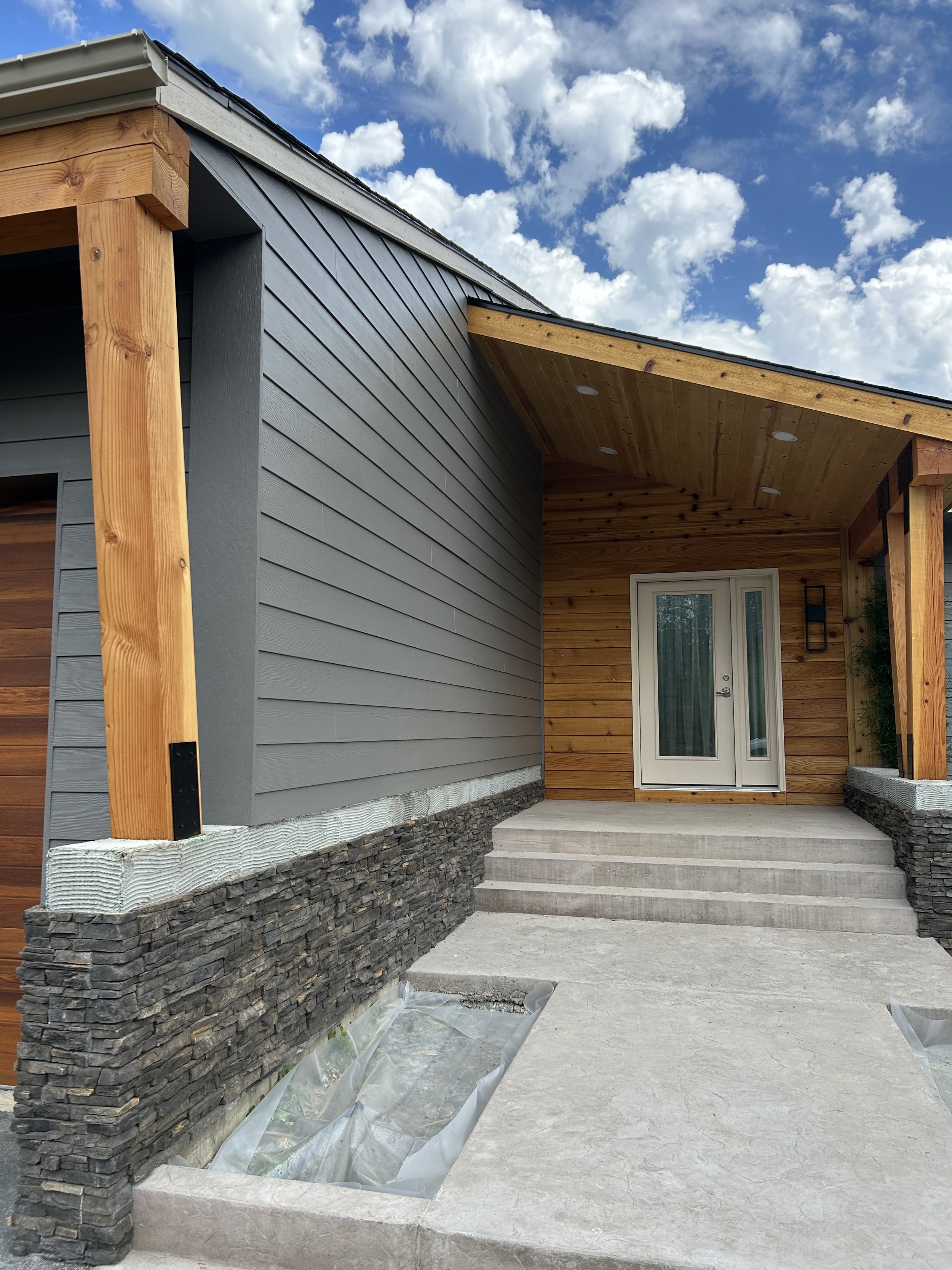 Newly constructed house entrance with concrete steps, stone veneer, and wooden siding under a blue sky with clouds.