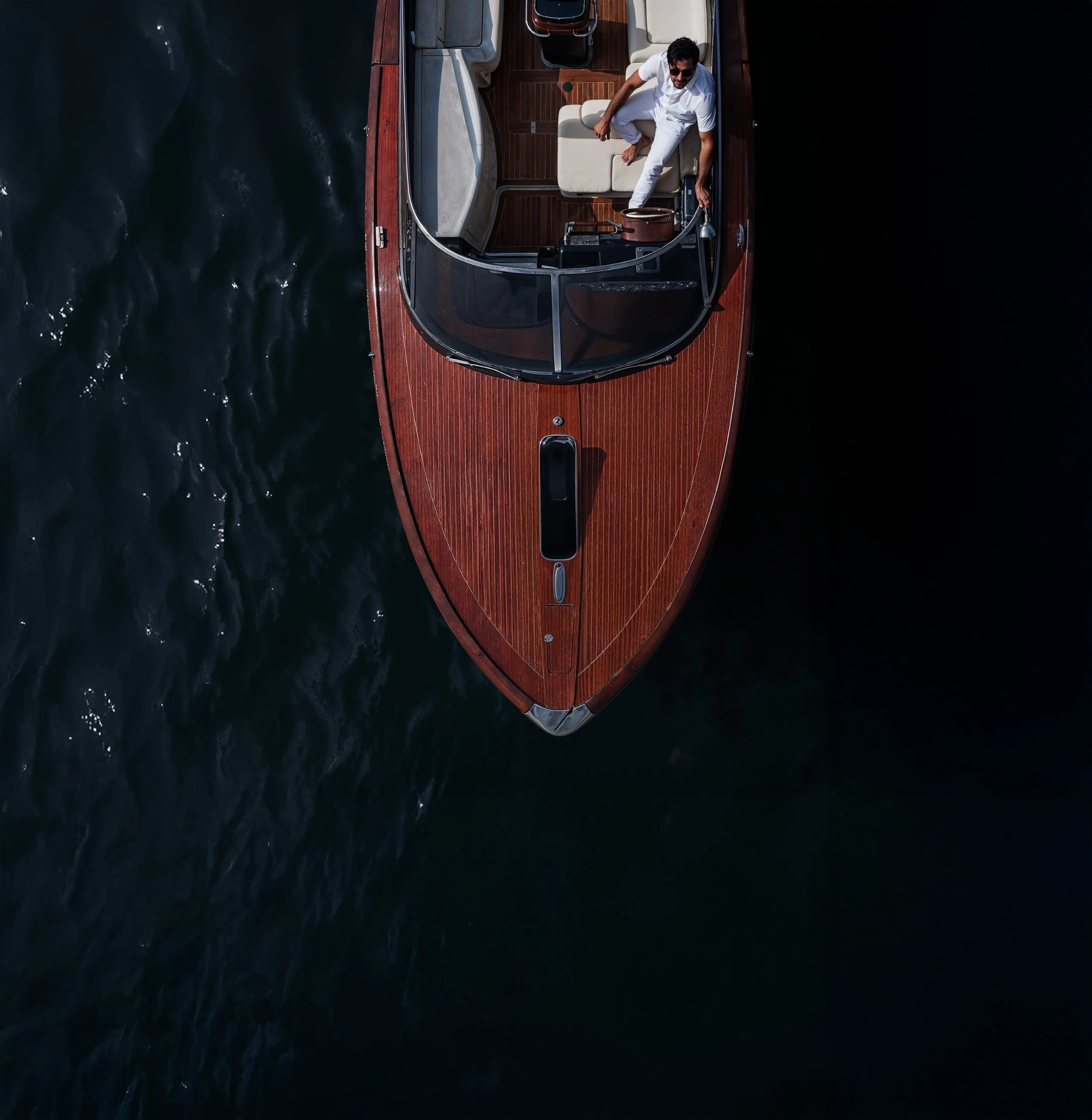A man with dark hair and a beard, wearing sunglasses and a white shirt, stands on the deck of a wooden boat, adjusting equipment near the railing. The boat has a polished wood surface, a white cushioned seat, and is on dark water.