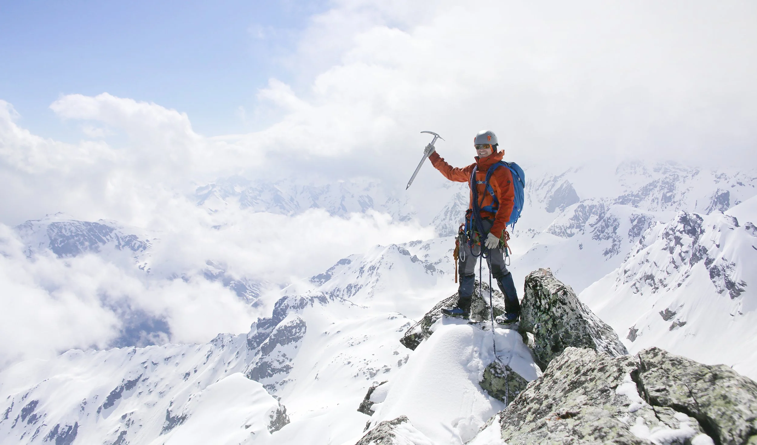 A person wearing bright orange mountaineering gear, including a helmet and gloves, standing on snowy rocky terrain in a mountain range. They are holding an ice axe in one hand and smiling with a backpack on. Snow-covered peaks and a cloudy sky are in the background.