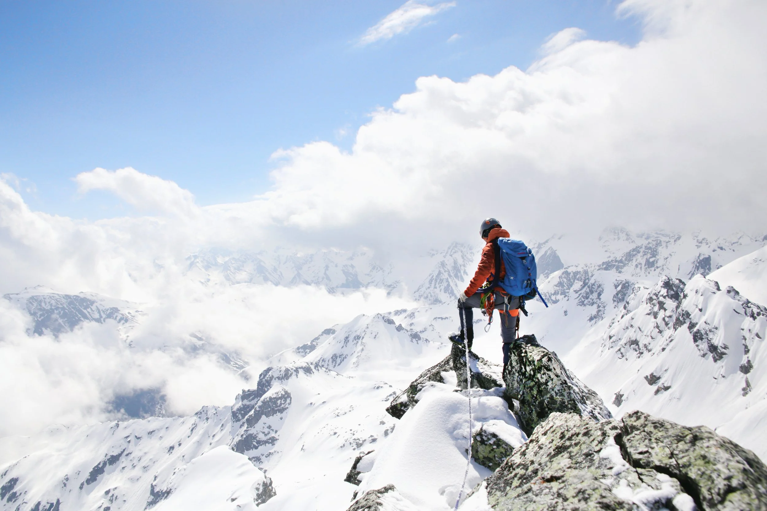 A person wearing winter gear, including a helmet, orange jacket, and blue backpack, standing on snowy rocks on a mountain, with snow-covered peaks and a partly cloudy sky in the background.