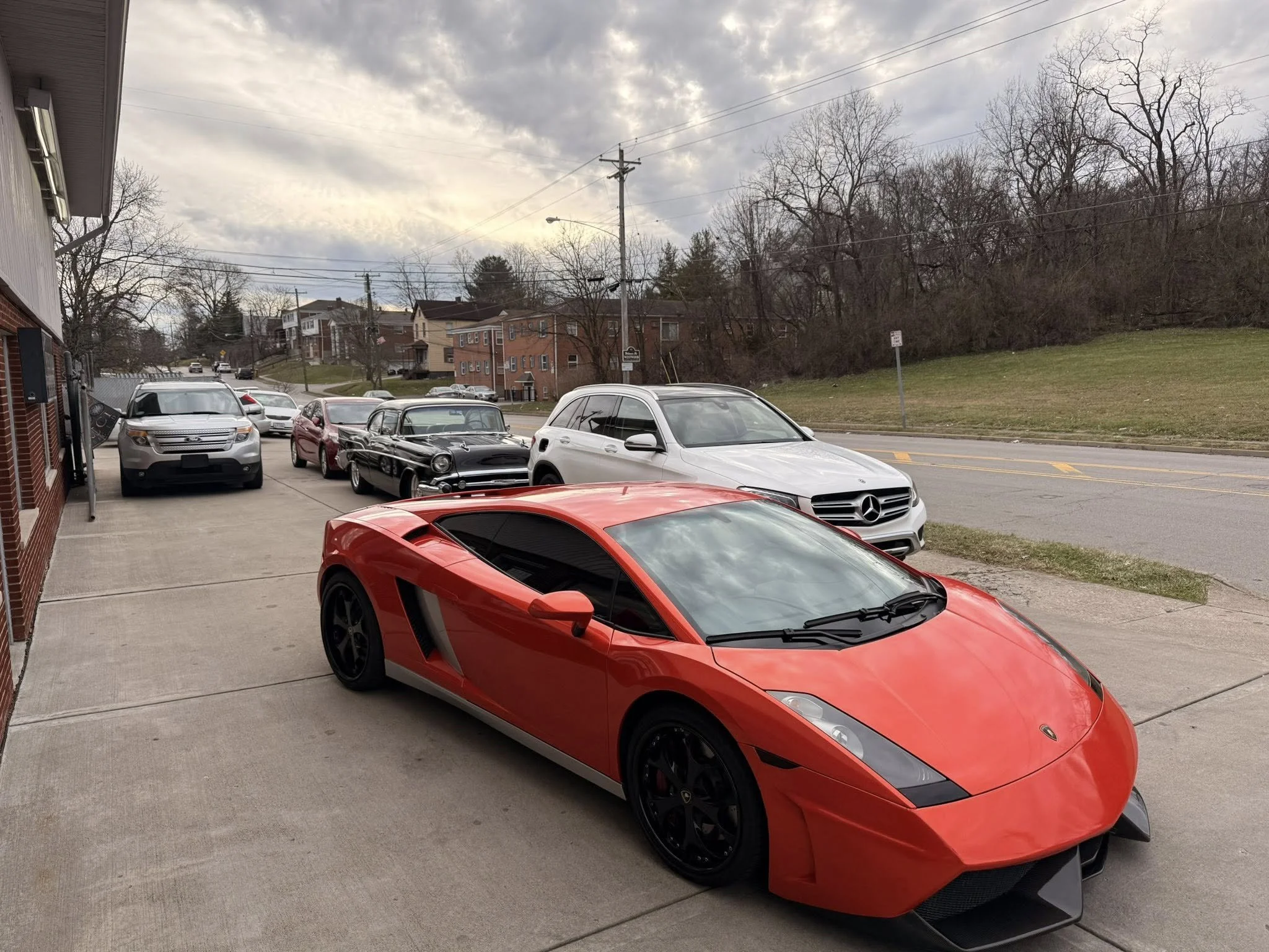 Red Lamborghini sports car parked in front of a building with several other cars behind it, including a white sedan, a black vintage car, and a gray SUV, with a residential neighborhood and cloudy sky in the background.