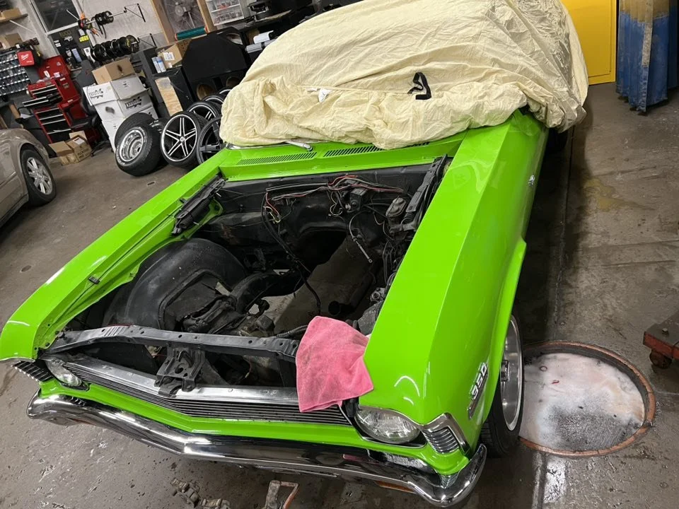 A bright green classic car with its hood removed, exposing the engine bay, partially covered with a beige protective sheet, inside a garage with tools and spare tires in the background.