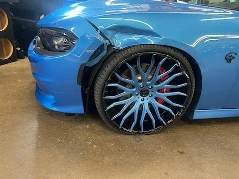 Blue sports car with a damaged front bumper and a large, decorative aftermarket wheel with a black and silver design.