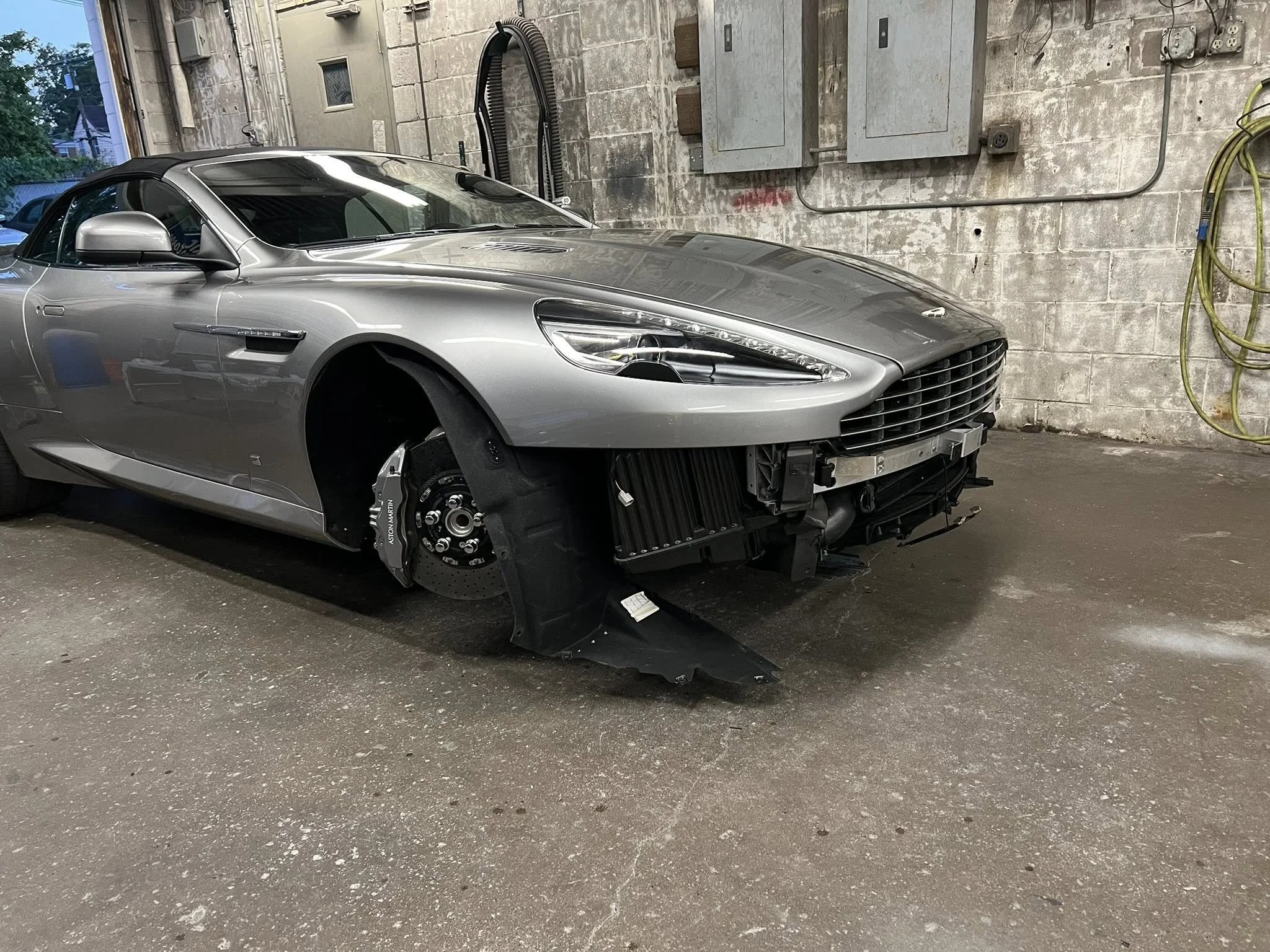 A silver sports car with front left wheel removed, parked inside a garage with a concrete wall and cleaning equipment.