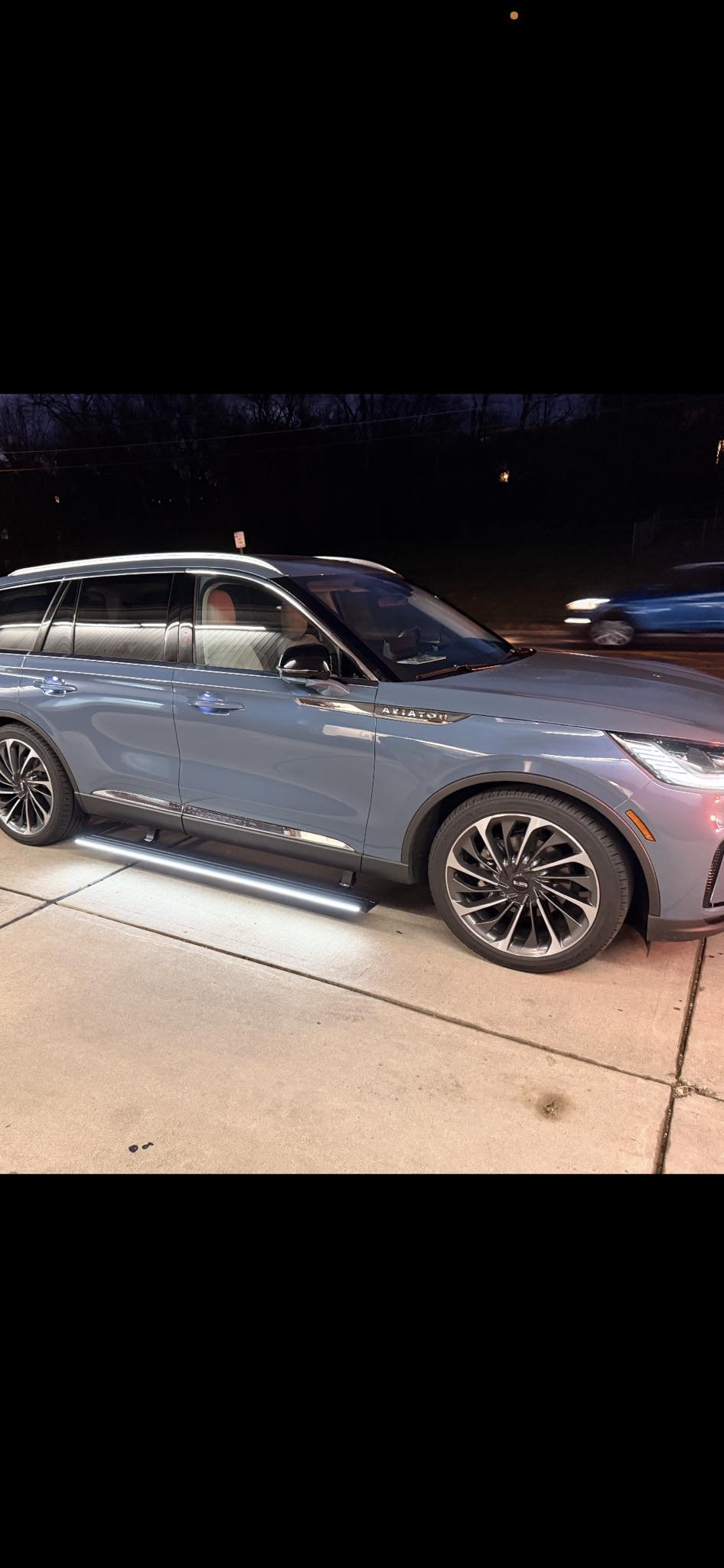 A blue Audi SUV parked on a concrete sidewalk at night, illuminated by nearby lights, with a car passing by in the background.