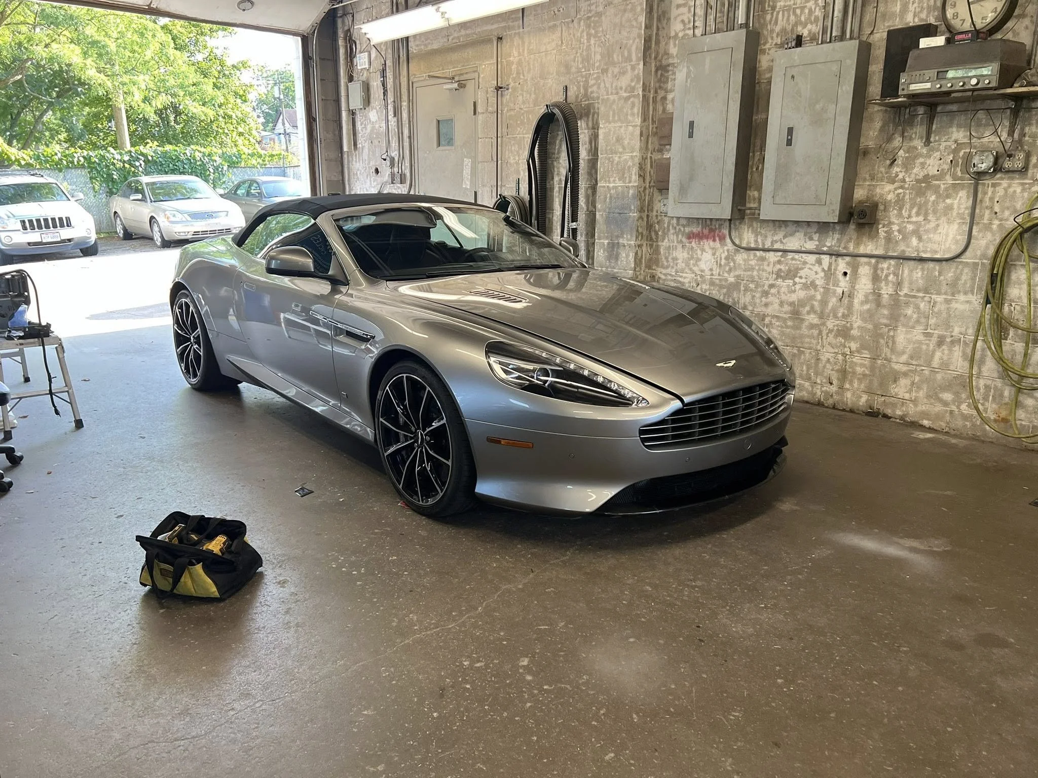 Silver convertible sports car parked inside a garage with an open door. There are three other cars visible outside through the garage opening. The garage has exposed brick walls, electrical panels, and hoses hanging on the wall.
