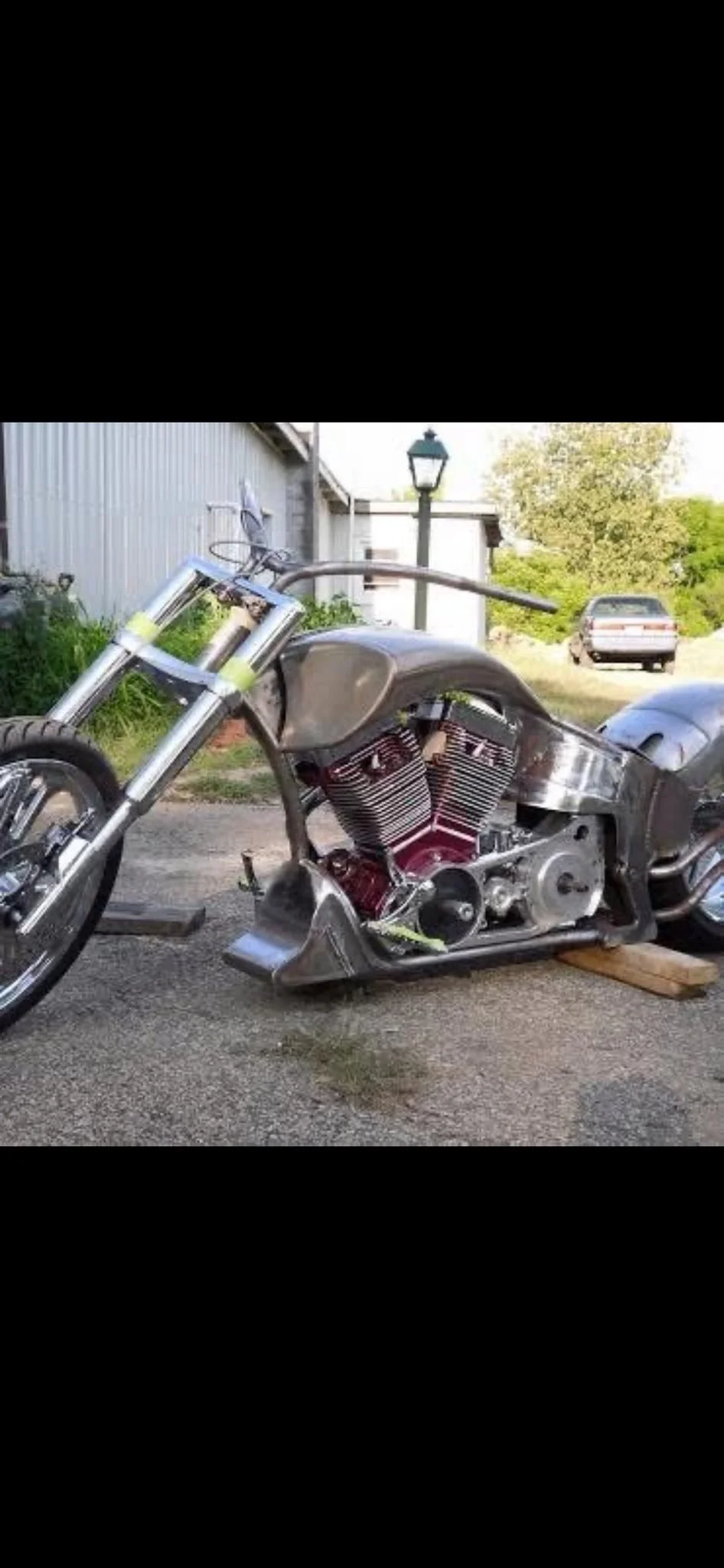 A motorcycle in the process of being built or repaired, with the frame and engine visible but unfinished, parked outdoors on a concrete surface near a shed and trees.