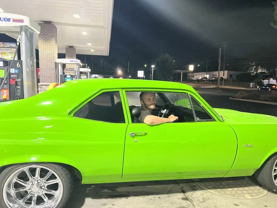 A bright green vintage car parked at a gas station at night, with a man inside smiling and looking at the camera.