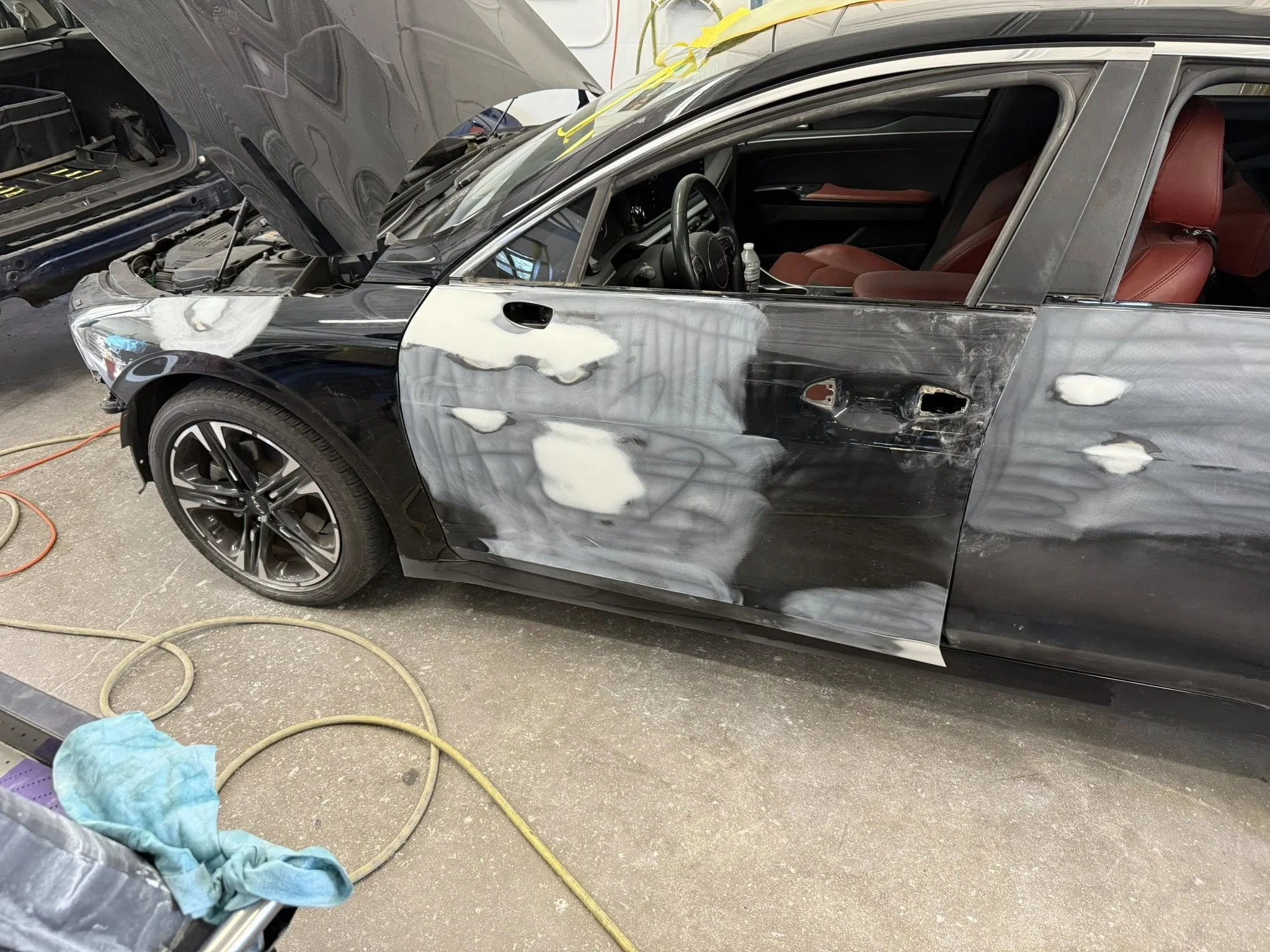 Black sports car undergoing body repair with sanded primer patches on the door and front fender, a red interior, and a raised hood in a workshop.