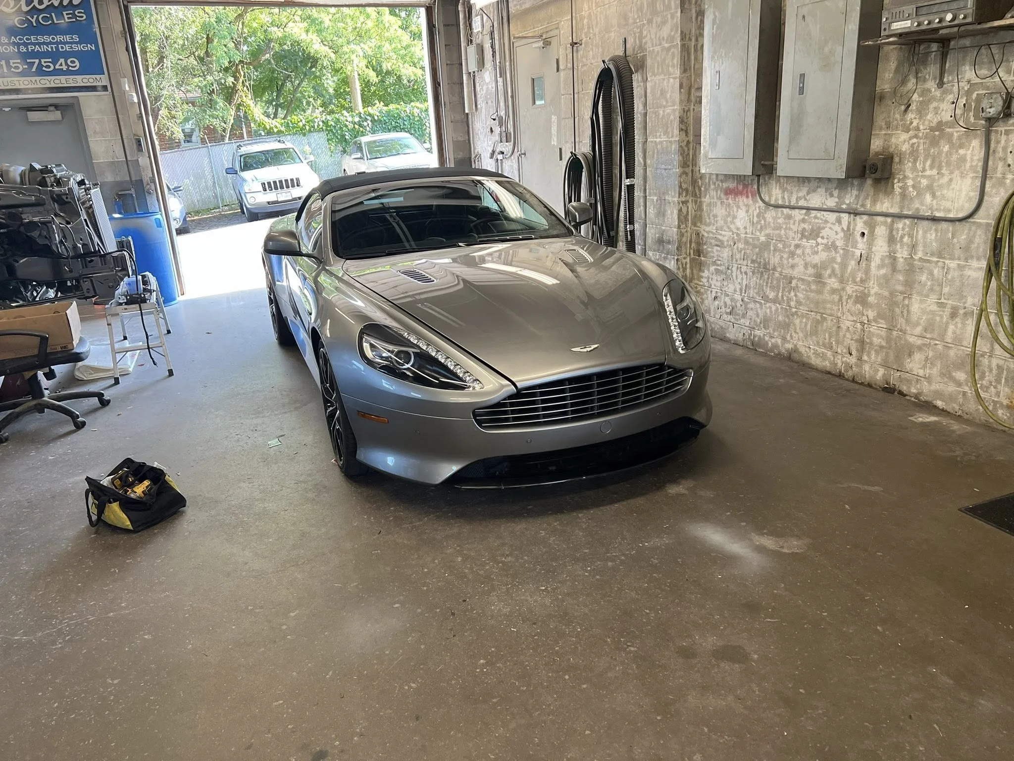 A silver Aston Martin sports car parked inside a garage with an open door, revealing trees and parked cars outside.