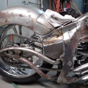 A close-up of a partially assembled motorcycle with a rusty exhaust pipe, a silver and rusty frame, and a white cloth draped over part of the tank, in a garage setting.