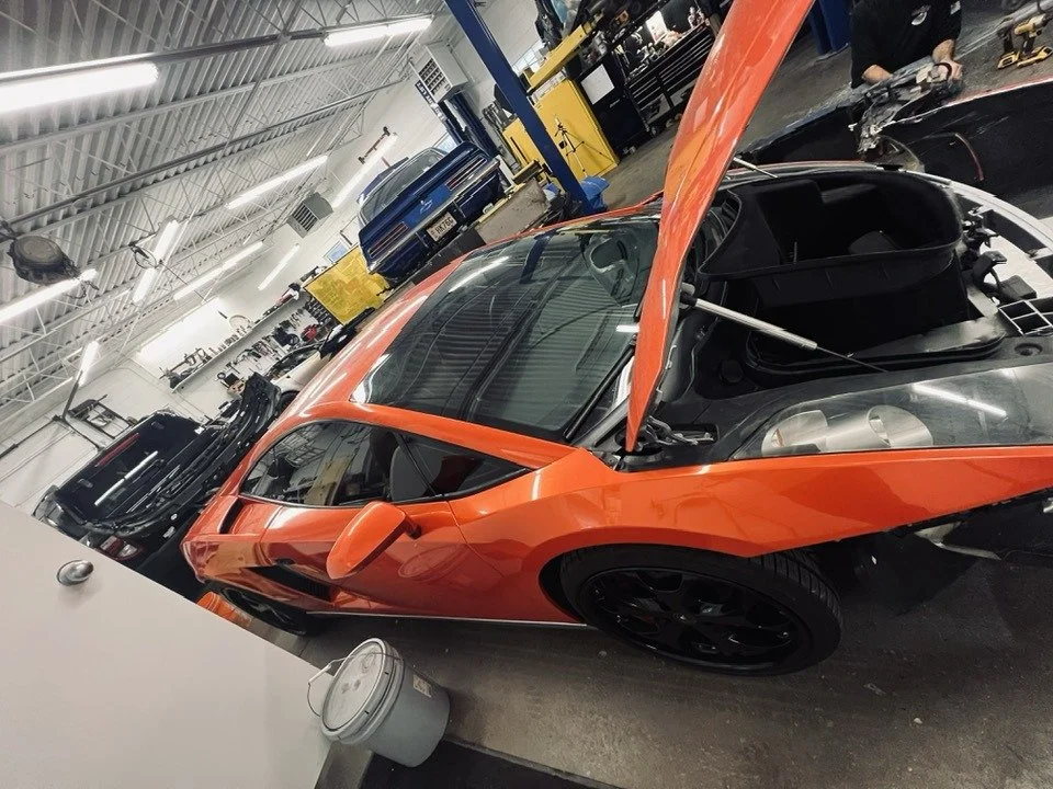 An orange sports car with its hood open inside an automotive workshop or garage.