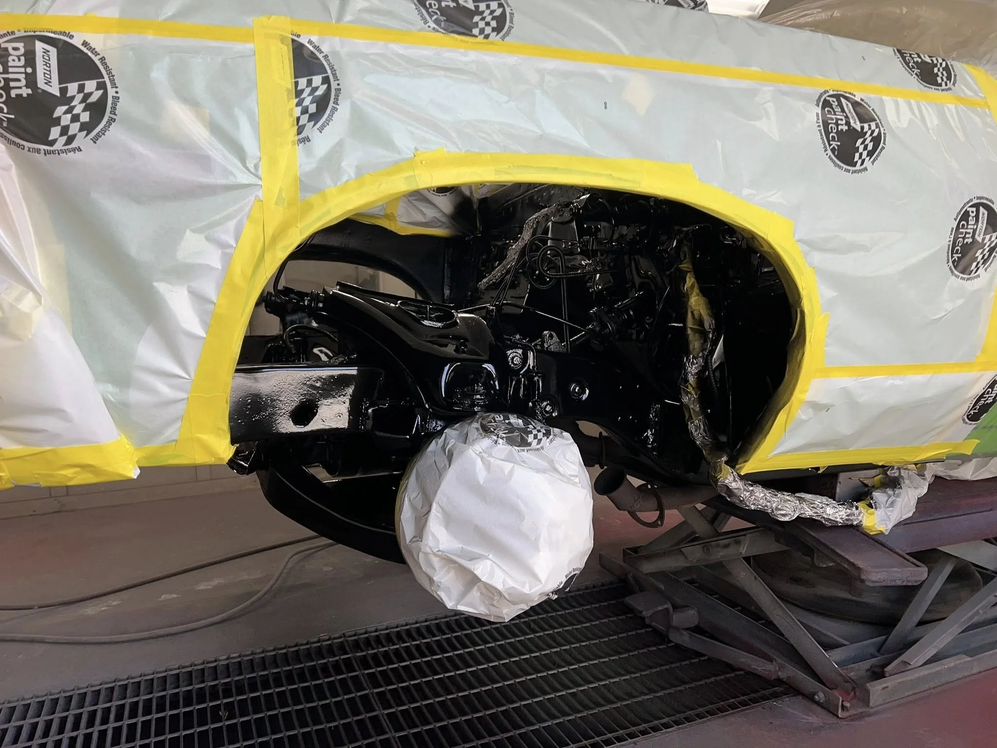 A motorcycle on a repair or painting stand, partially covered with masking paper and tape, possibly in a workshop or paint booth.