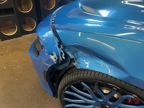 A blue sports car with front-end damage, including a scratched fender and missing panel, parked in a garage with wine bottles in the background.