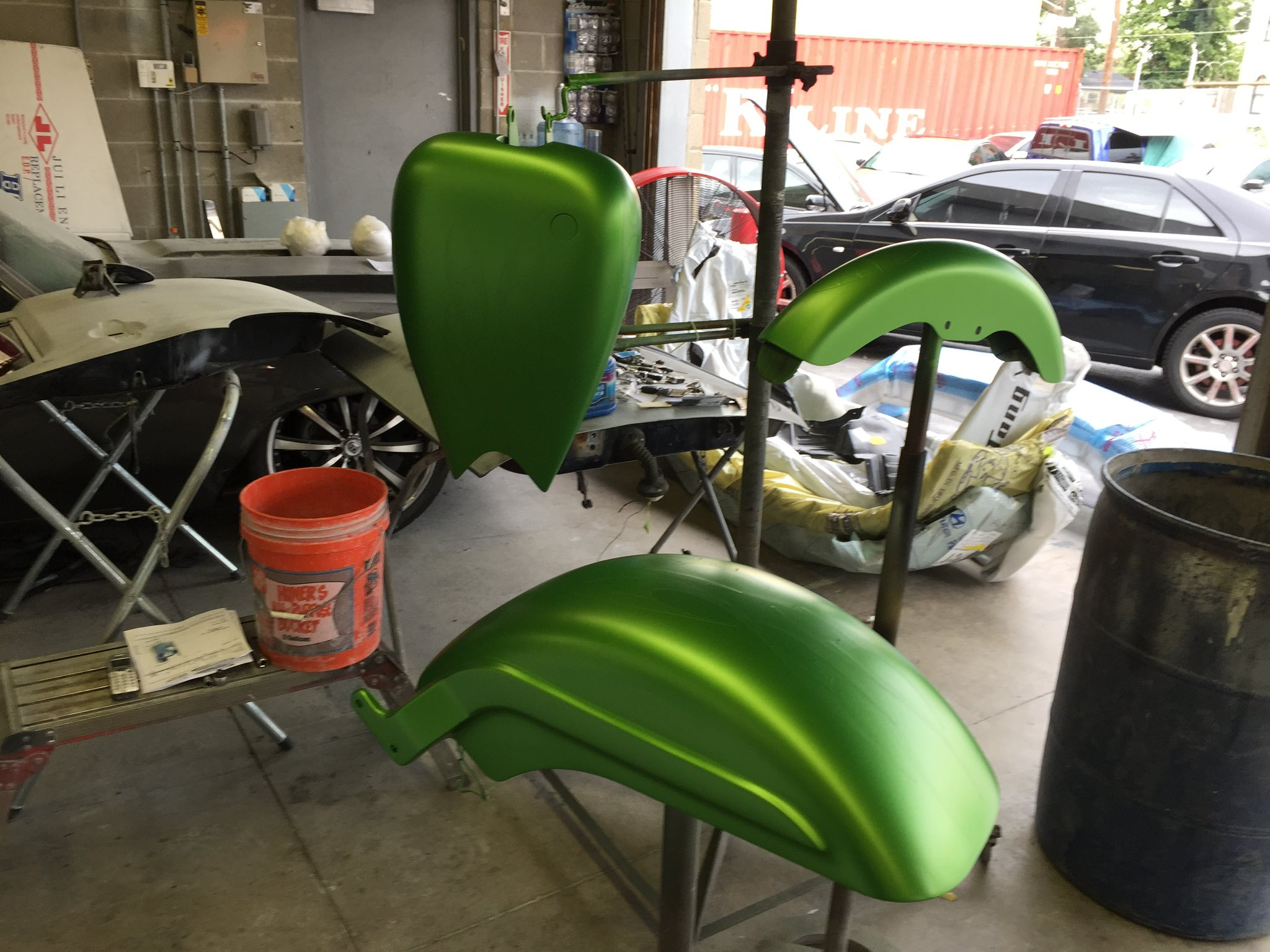Green motorcycle body parts hanging on a stand inside a workshop, with a car and various tools and supplies in the background.