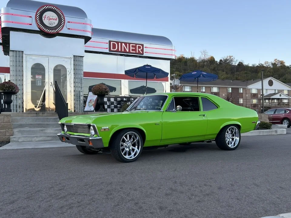 A bright green vintage muscle car parked outside a retro diner with a checkered awning and umbrellas, on a clear day with a few buildings and cars in the background.