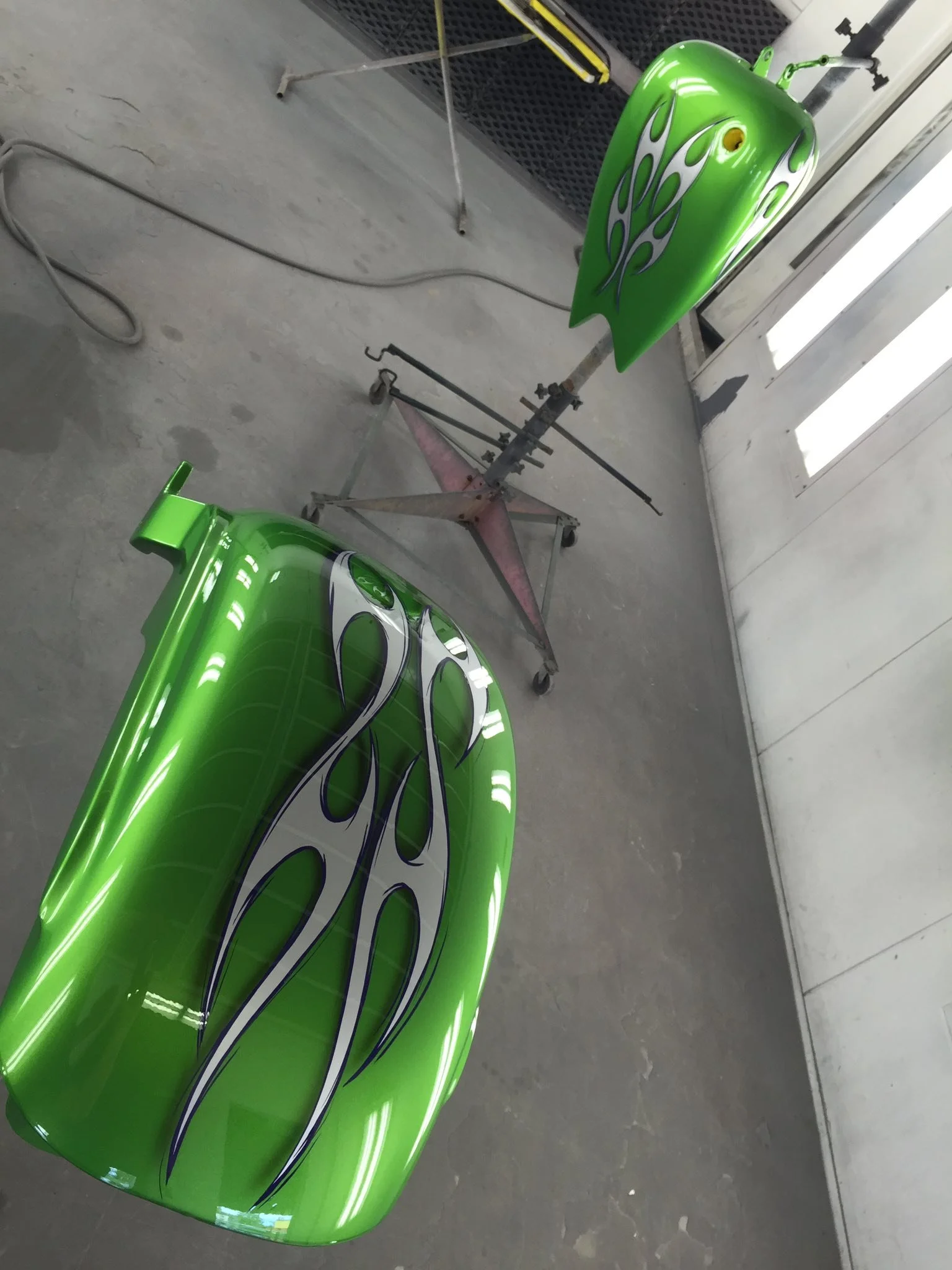 Two green painted motorcycle fairings with flame designs rest on a metal stand in a garage or workshop. The background shows a concrete floor and a wall with a window.