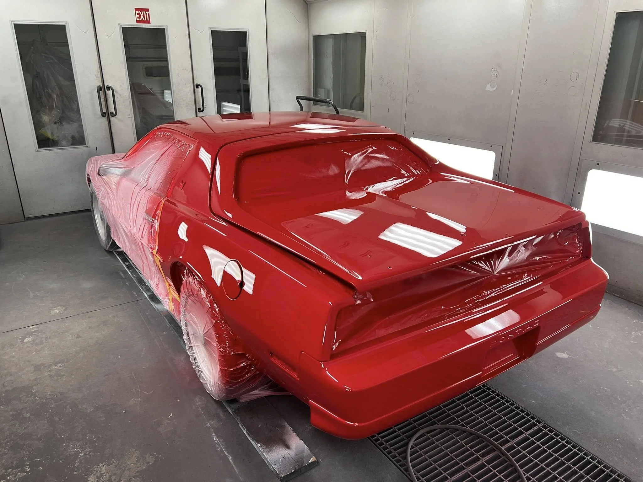 Red vintage sports car in a paint booth being painted, with plastic coverings on the wheels and windows, and bright work lights reflecting off its shiny surface.