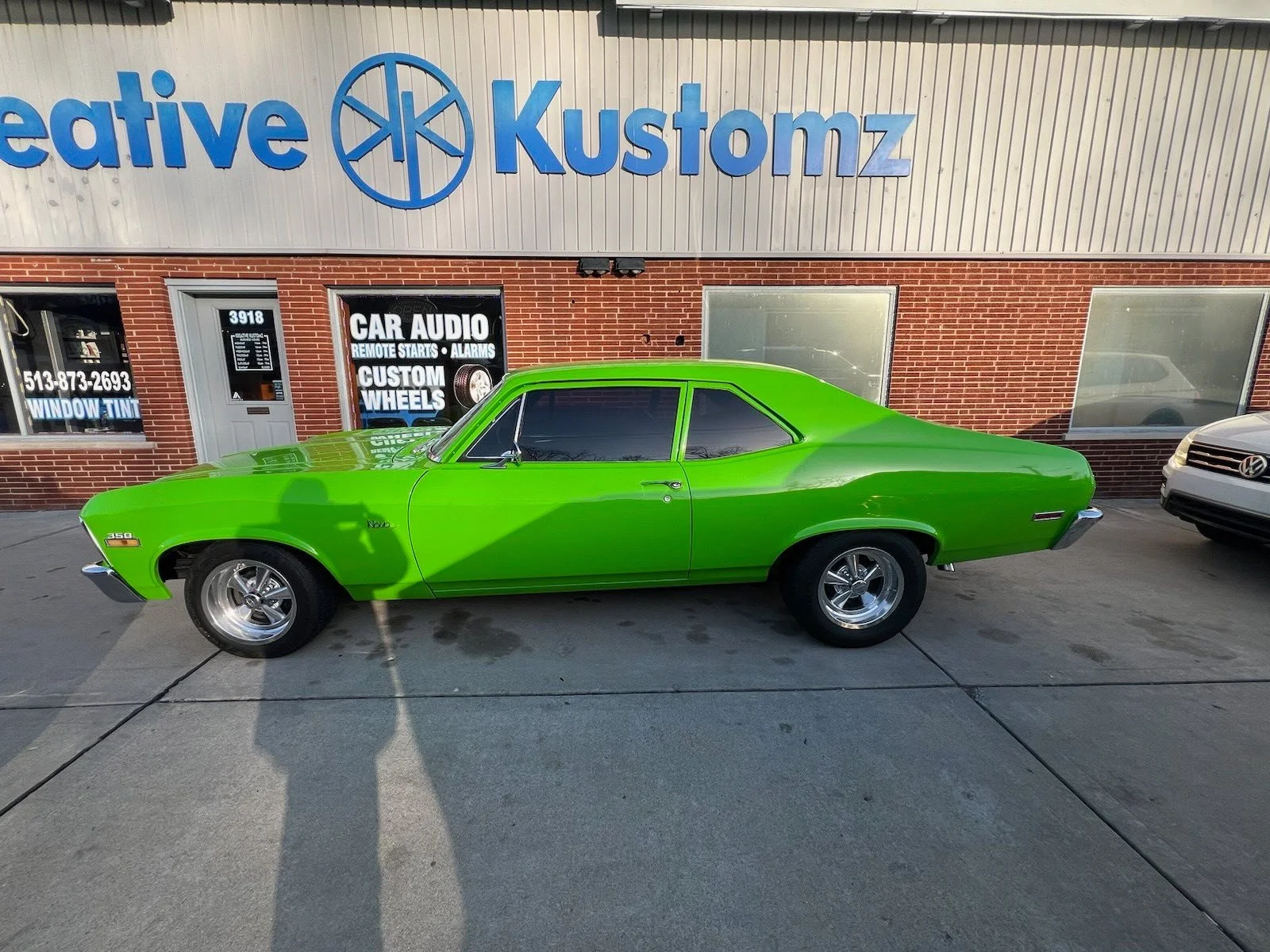 A bright green classic car parked outside a storefront with blue signage that reads 'Kustomz' and promotional signs for car audio, remote starts, alarms, custom wheels, and window tint.