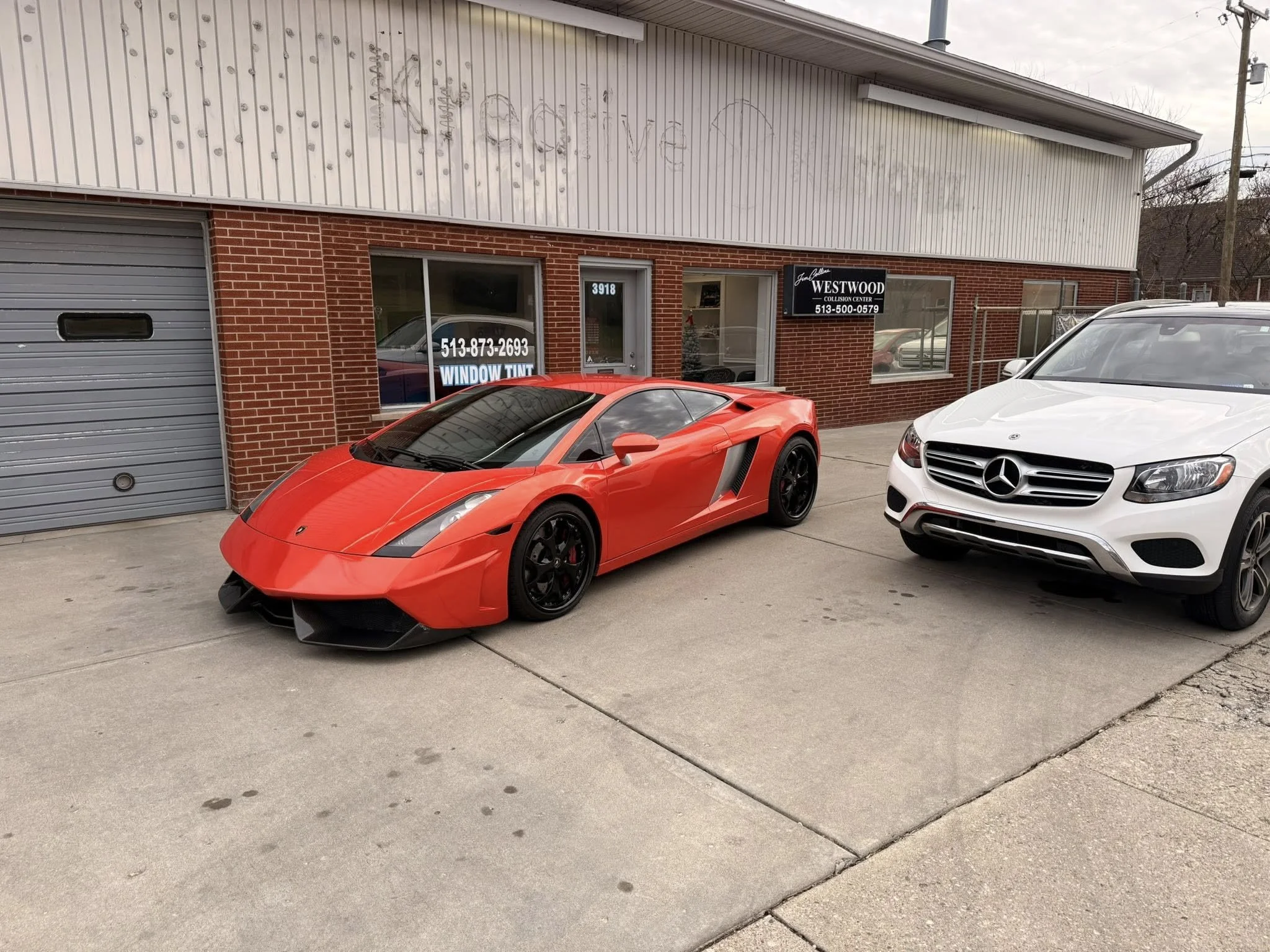 A red Lamborghini Gallardo parked on the street next to a white Mercedes-Benz GLK SUV in front of a brick building with a sign for a collision center and a window tinting service.
