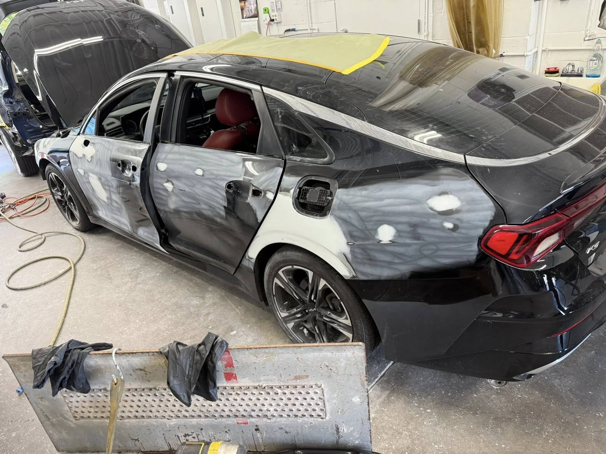 A black sedan car is in an auto body repair shop, with its passenger side door removed. The car has patches of primer and sanding marks on the side. There are tools, hoses, and a workbench with gloves and supplies nearby.