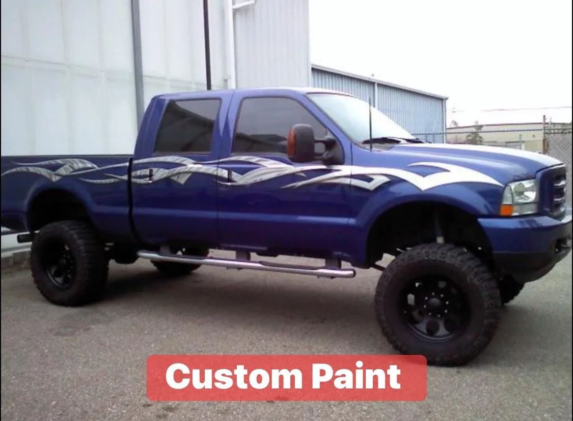 A large blue pickup truck with custom white and black paint designs on the hood, sides, and top, parked outside an industrial building. The truck has oversized tires and black rims.