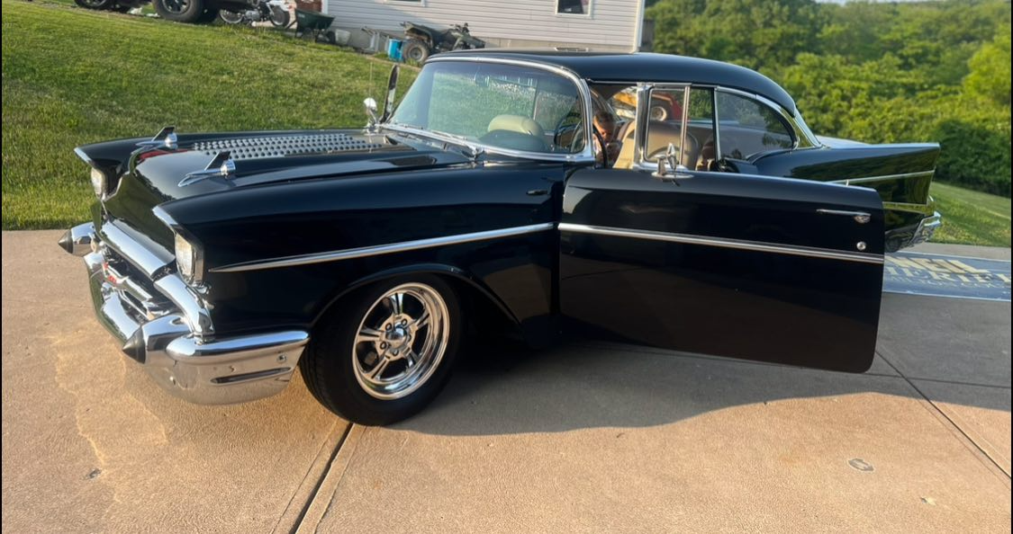 A classic black 1950s car parked on a driveway, with the driver's side door open, showing its vintage design and chrome details.