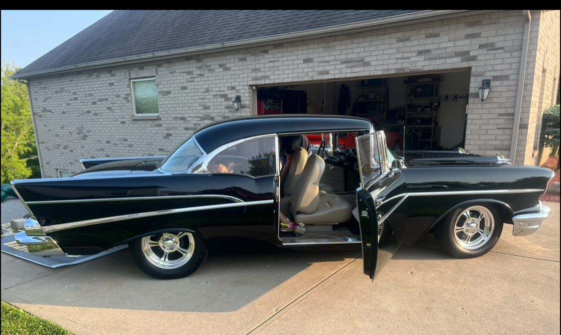 A classic black and white vintage car with its front opened door revealing a beige interior, parked on a driveway in front of a brick garage.