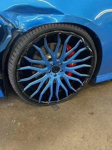 Close-up of a blue car's rear wheel with custom multi-spoke black and blue alloy rim and red brake caliper visible behind.