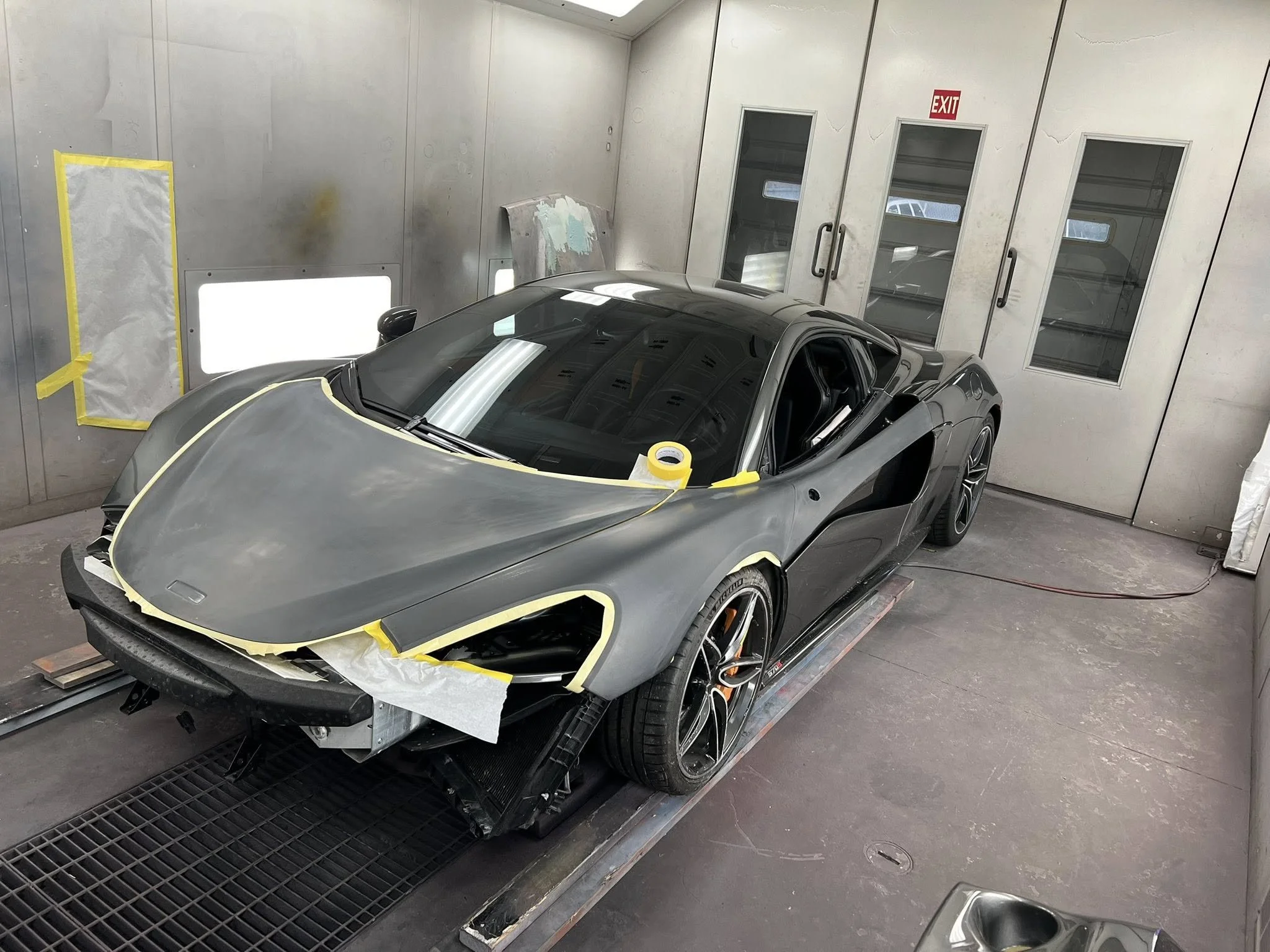A sleek black sports car inside a paint booth, partially masked for painting, with tape on the front and a cover on the windshield, surrounded by spray equipment and paint prep area.