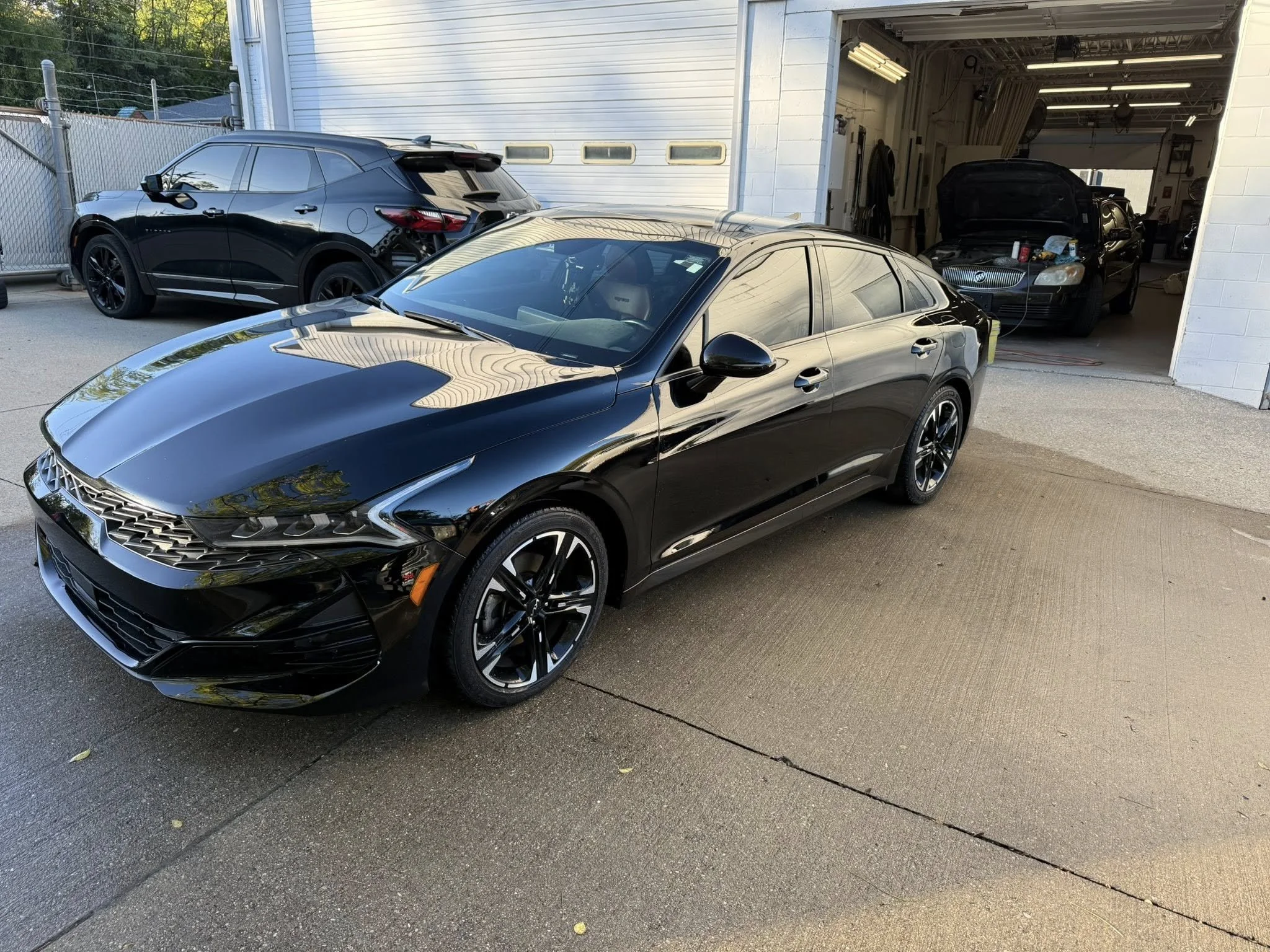 Black sedan parked outside a garage, with other cars inside and outside the garage.