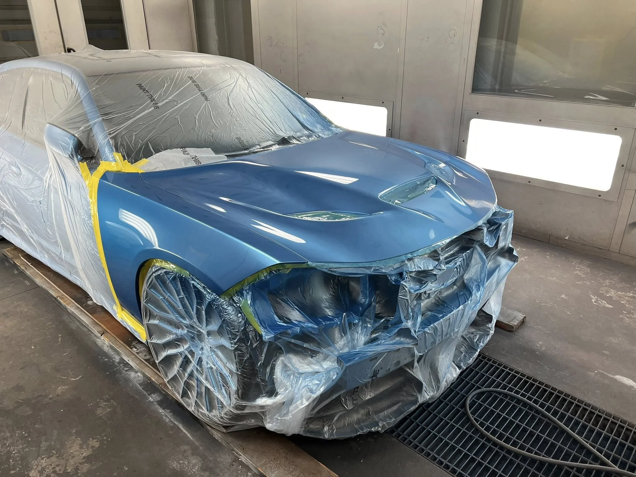 A blue sports car being painted in a spray booth, covered in protective plastic and masking tape to shield areas from paint.
