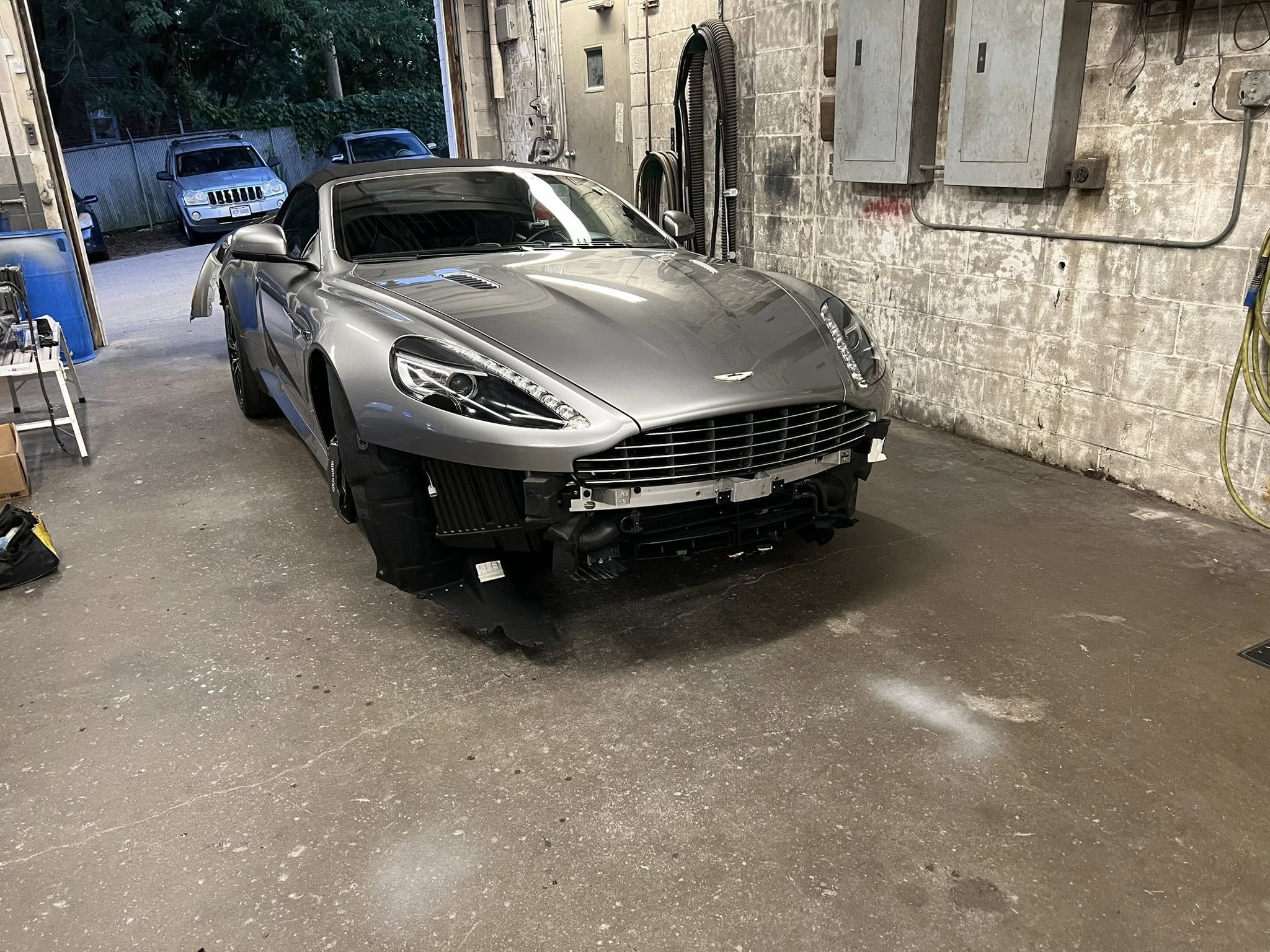 A silver sports car, likely an Aston Martin, is inside a garage with part of the front bumper removed, exposing internal components.