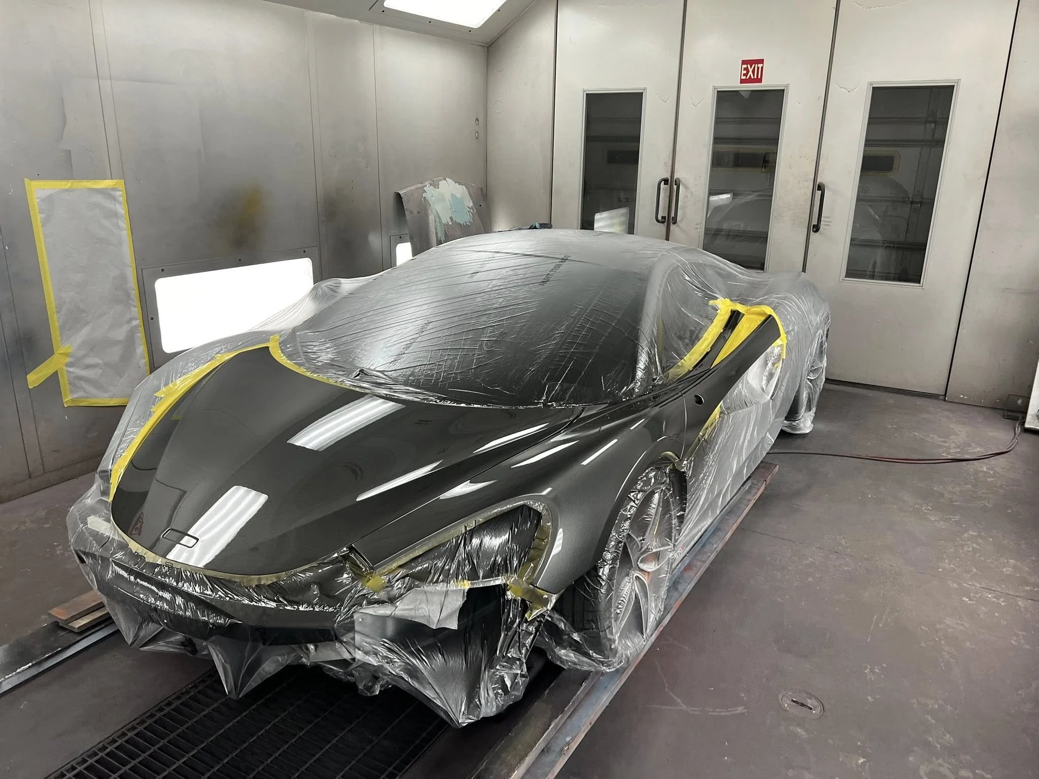A sleek black sports car inside a paint booth, partially covered with protective plastic. The room has metallic walls, an exit door with glass panels, and bright overhead lights.