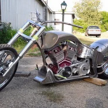 An unfinished or custom motorcycle parked outside on a driveway with a building, a car, trees, and a lamp post in the background.