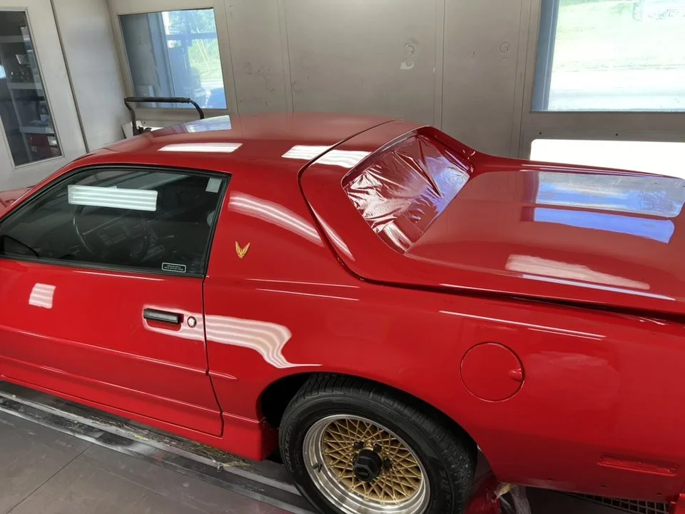 A red sports car parked indoors near windows, with a small V-shaped emblem on the driver's door and gold mesh wheels.