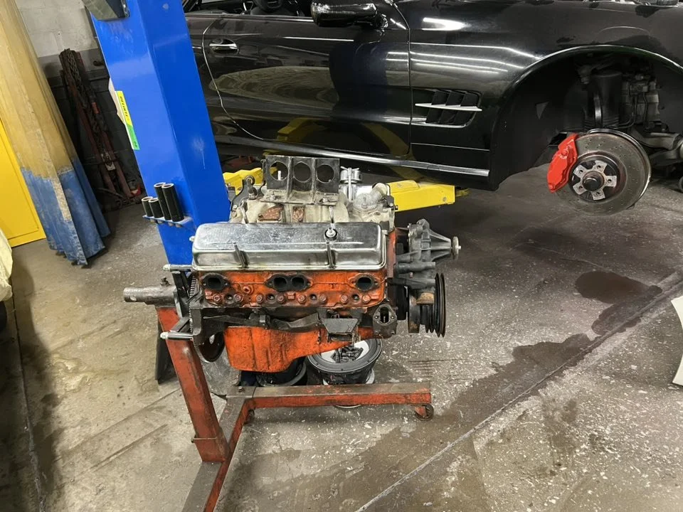 An automotive repair shop with a black sports car on a lift and a disassembled engine on a stand in the foreground.
