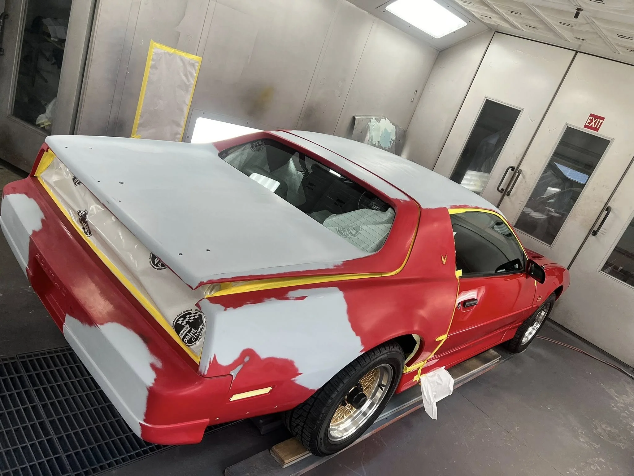 A red sports car in a paint booth undergoing a repaint or restoration, with parts masked and primed areas visible.