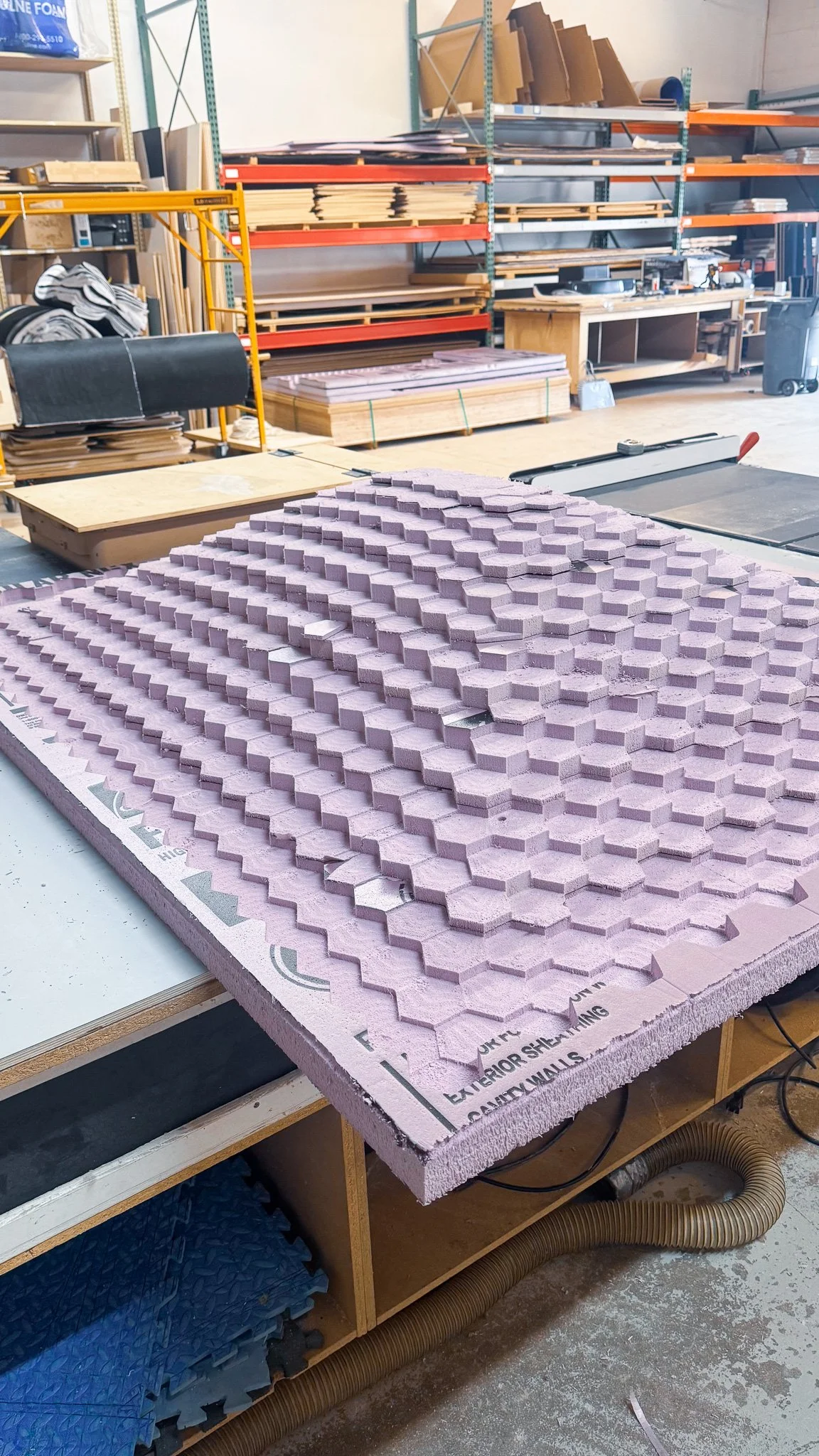 A workbench with pink foam insulation cut into a honeycomb pattern in a woodworking or construction shop. Shelves stocked with wood pieces, panels, and tools are visible in the background.