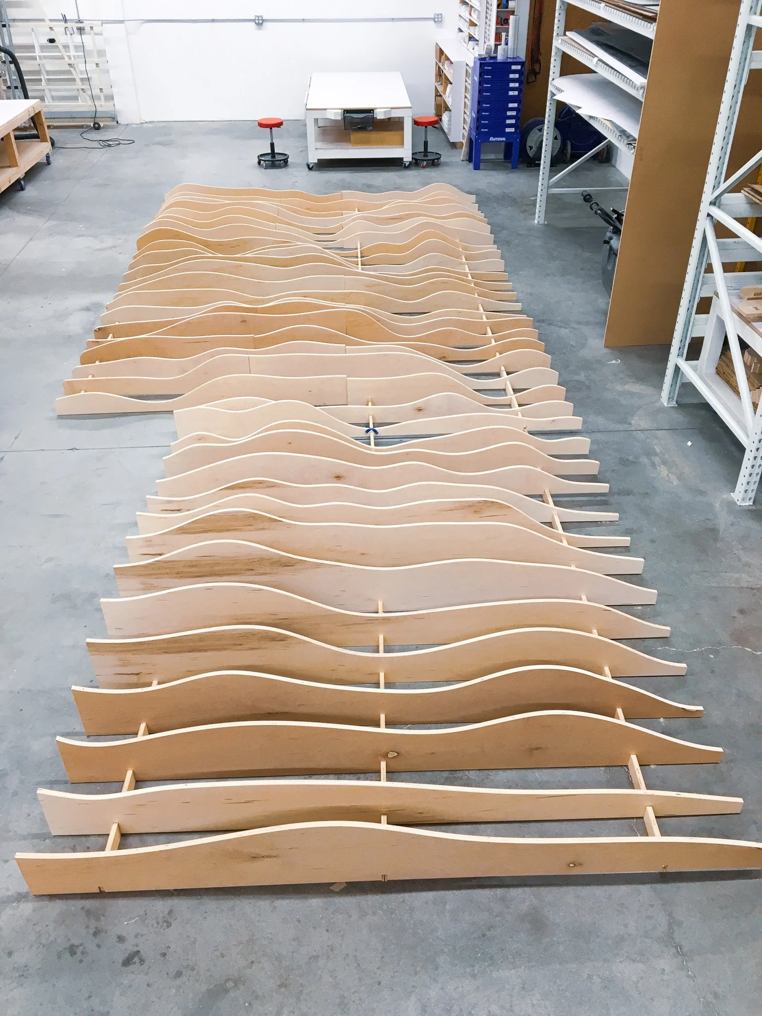 Set of wooden furniture pieces with curved sides, possibly parts for a bed or shelving, arranged on the floor of a workshop or storage area with shelves and tools in the background.