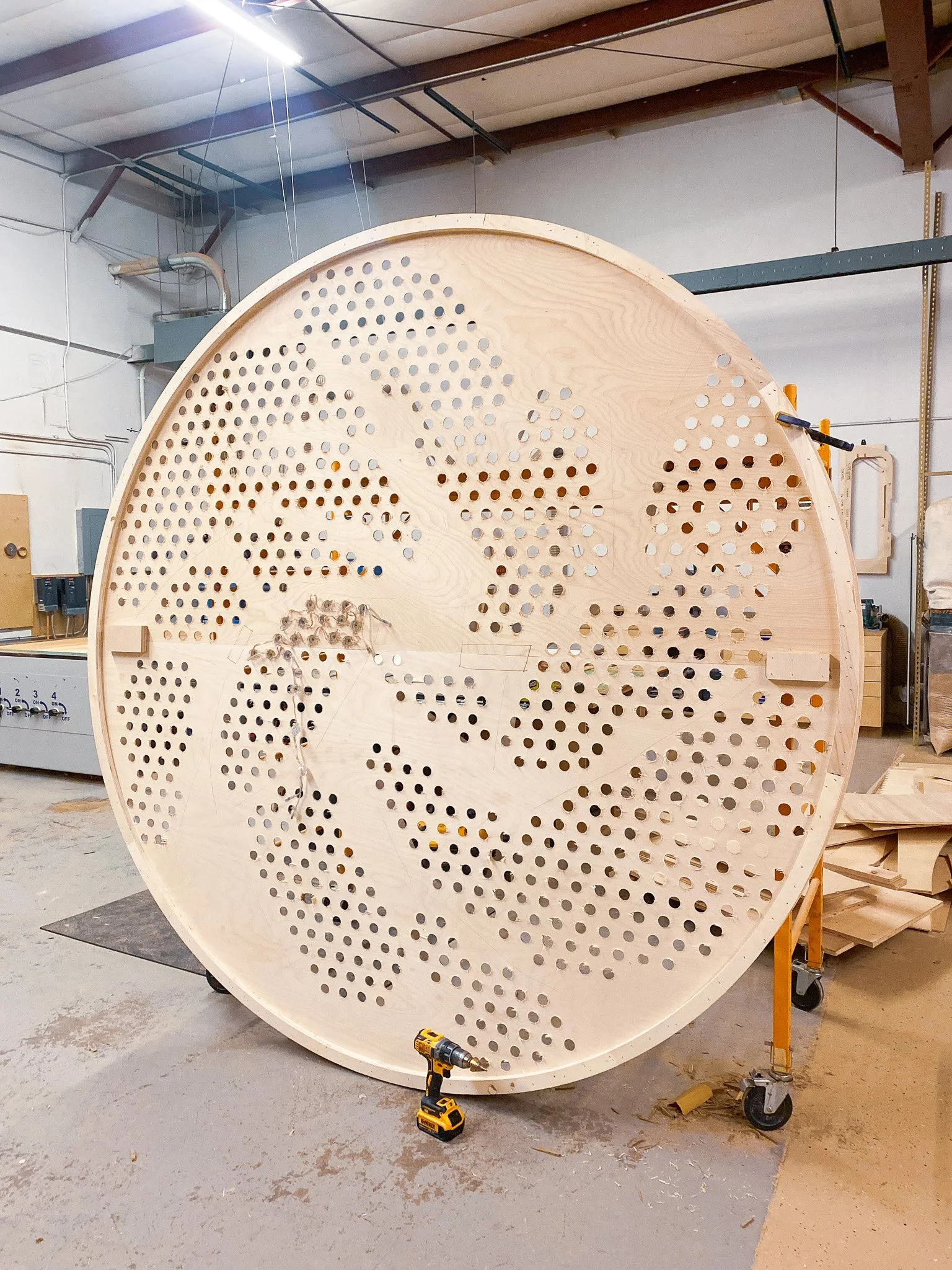 Large circular wooden structure with multiple small holes drilled into its surface, resting on a workbench in a wood workshop. Various tools and construction materials are visible around.
