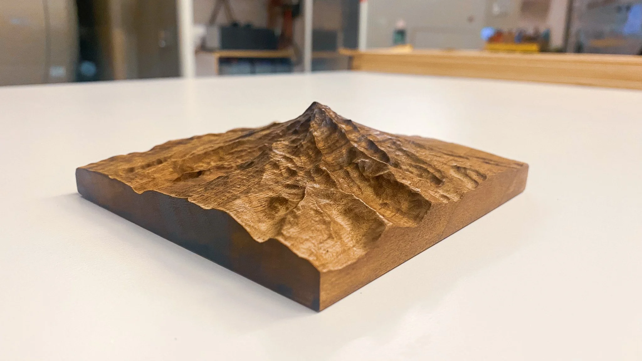 A small carved wooden mountain on a rectangular wooden base, with a textured carved surface and a peak in the center, placed on a white surface.