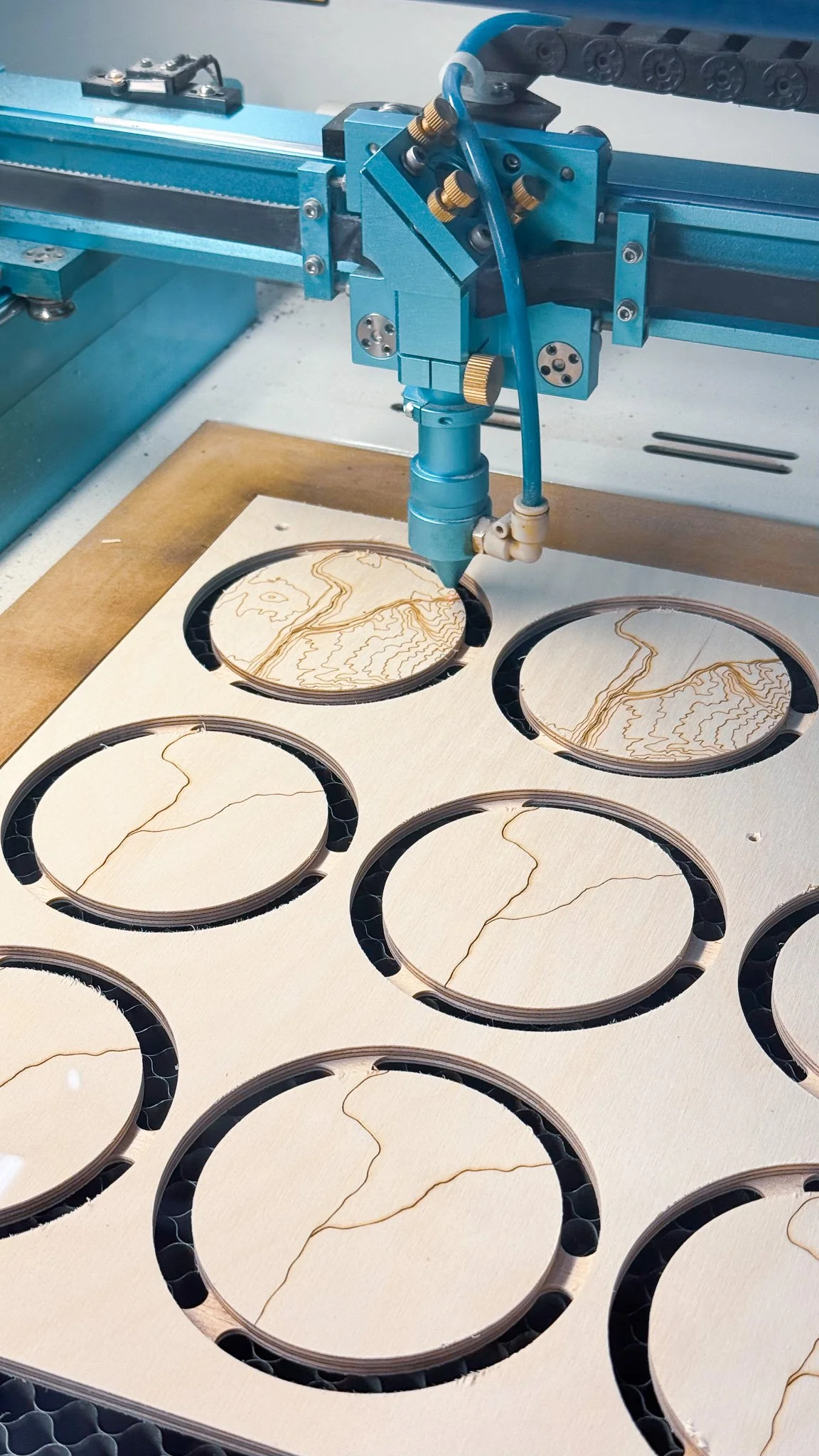 A laser cutter engraving designs onto wooden circular pieces, arranged on a cutout board.