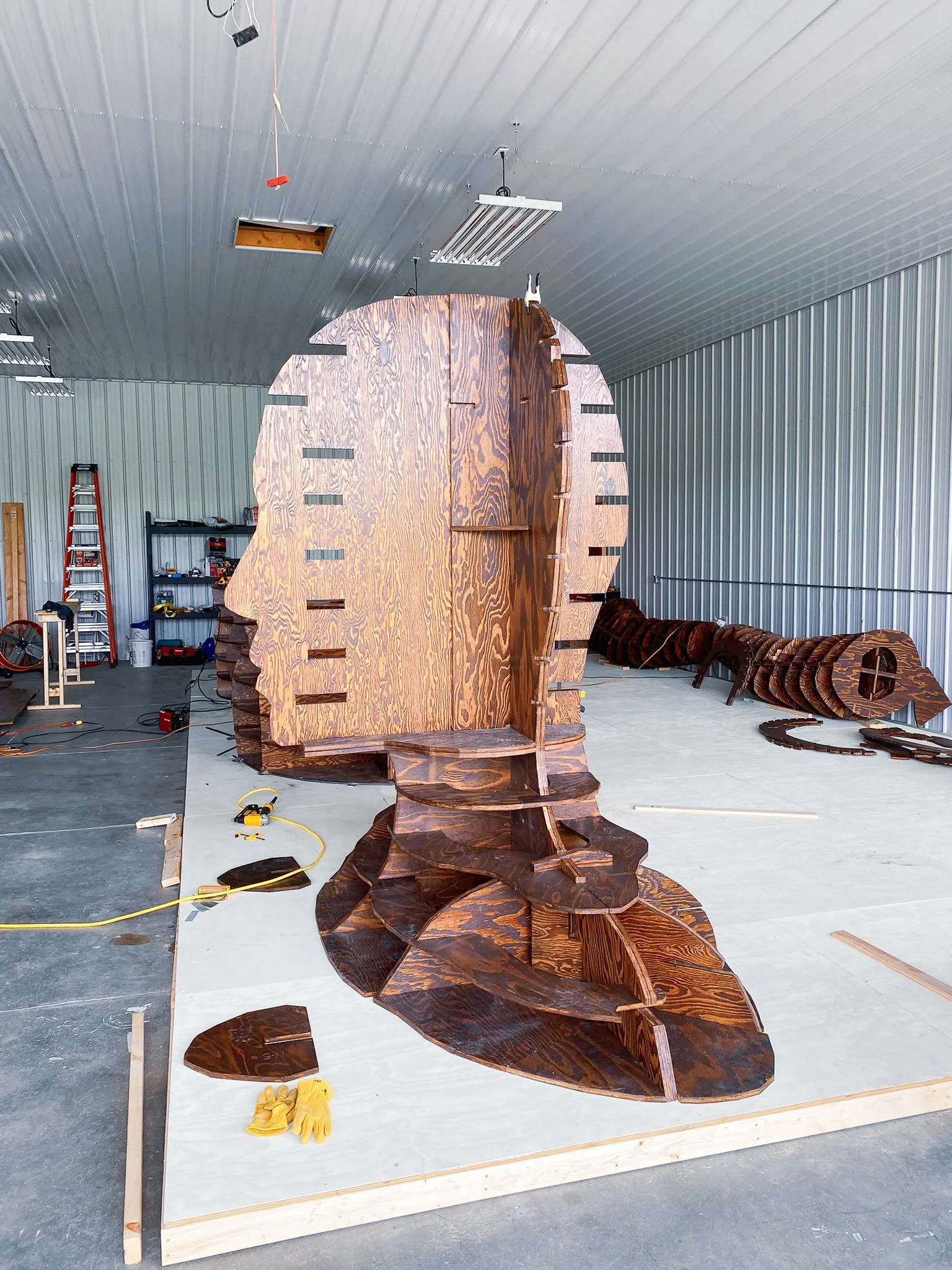 A large wooden structure being built inside a workshop, with multiple curved wooden pieces and a metal work surface, several tools, and a ladder in the background.